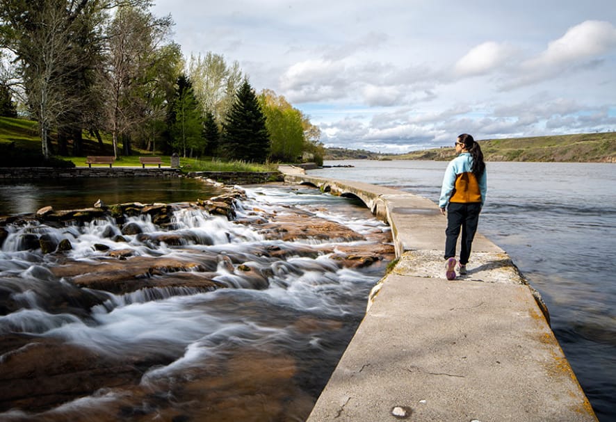 Student wearing backpack and walking on path next to water and small waterfalls.